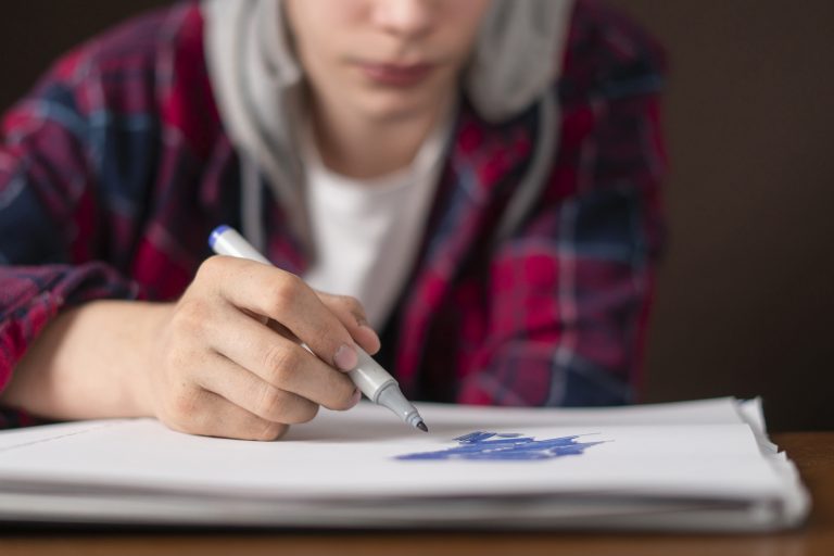 young male sitting at the table and drawing pictures in his notebook with marker b