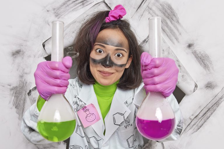 Female laboratory researcher holds two flasks with colorful liquid has dirty face after explosion wears medical coat and gloves shows results of her experiment in lab breaks through paper hole